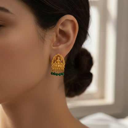 Gold plated earring with Lakshmi motif and green gemstones worn by a person, blurred background