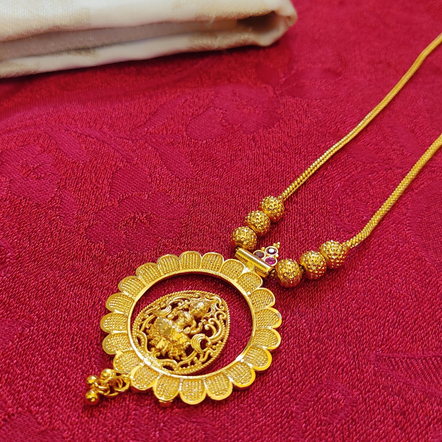 Gold plated necklace with intricate design and matte finish lakshmi on a lotus shaped central pendant on a red fabric background