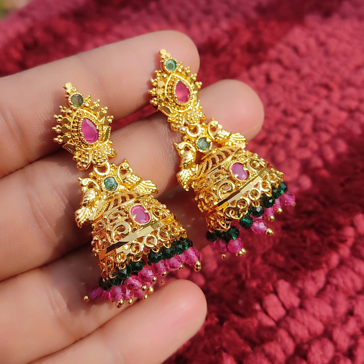 Gold plated Peacock motif Jhumka/jimikki earrings with pink and green stones held between fingers against a red textured background