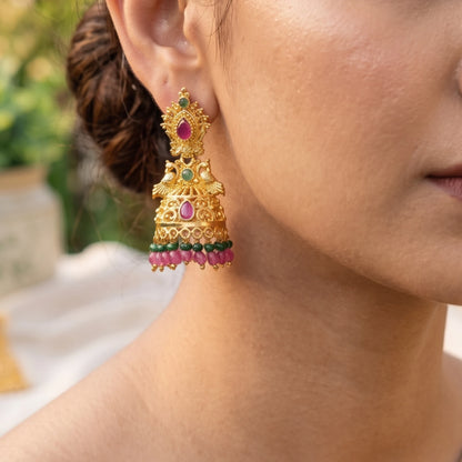 Gold plated Peacock Motif Jhumka/Jimikki earrings with pink and green stones worn by a person.