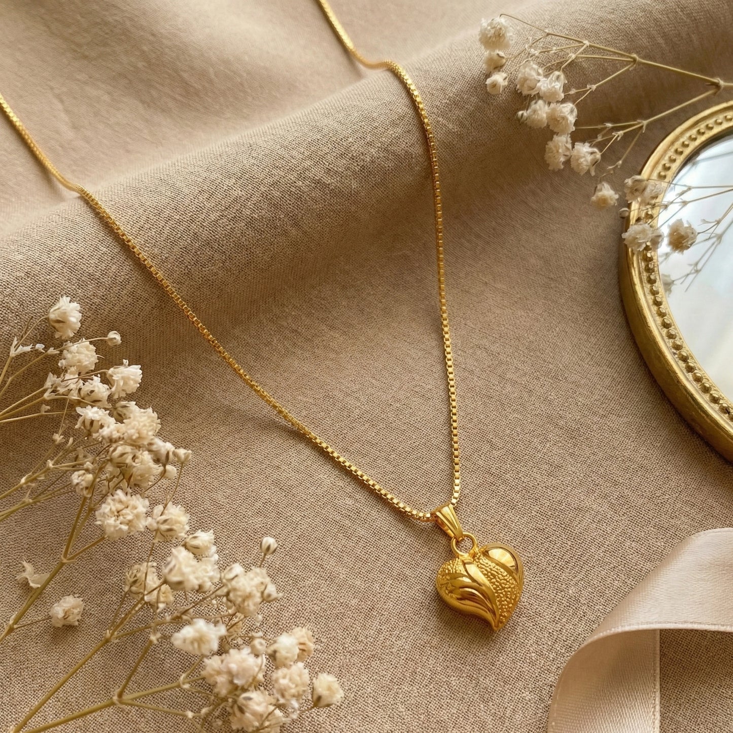Gold plated neckchain with a heart pendant on a beige fabric background with dried flowers.