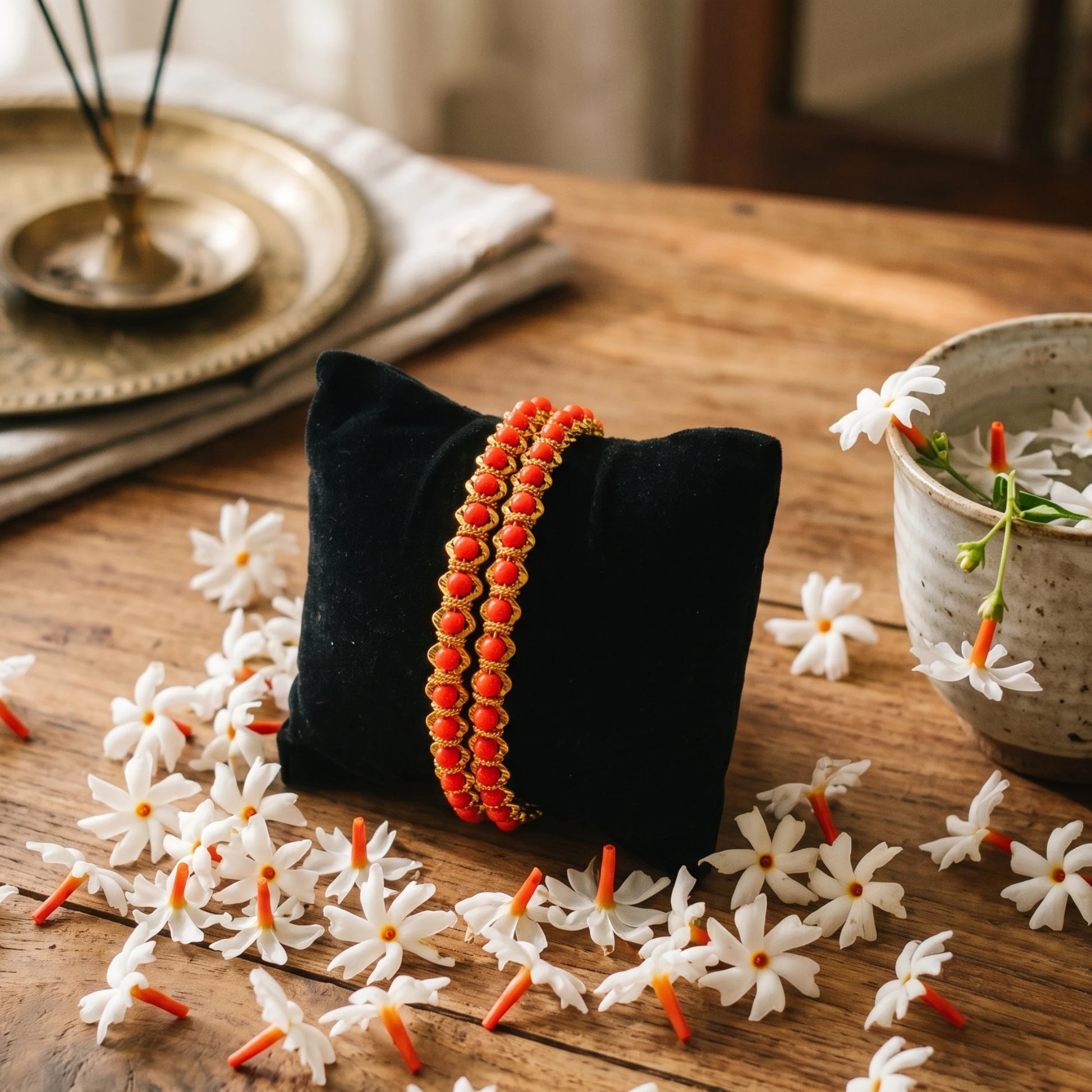 A pair of gold plated red coral beaded bangles displayed on a black velvet pillow, set on a rustic wooden table surrounded by white flowers.