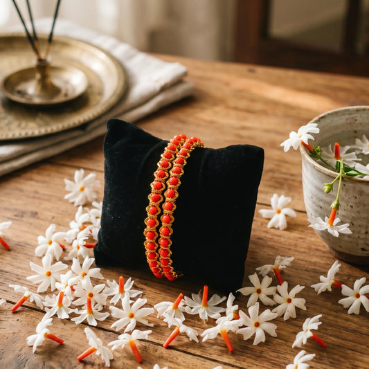 A pair of gold plated red coral beaded bangles displayed on a black velvet pillow, set on a rustic wooden table surrounded by white flowers.