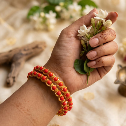 A close-up of a person's hand and wrist wearing two gold plated red coral beaded bangles, holding a small bunch of white flowers against a soft beige fabric background.