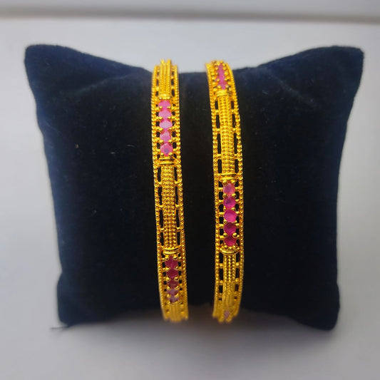 Two gold plated bangles with pink stones and filigree work on a dark blue cushion