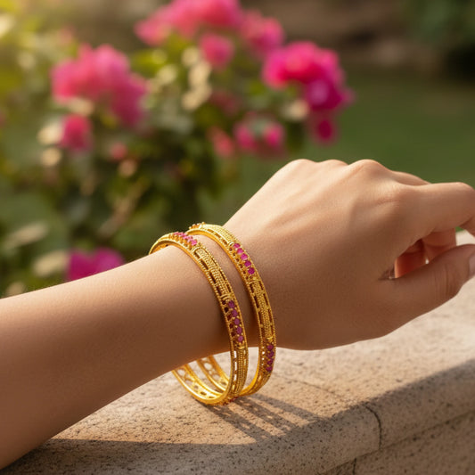 Hand wearing gold plated bangles with pink stones and filigree work against a blurred floral background