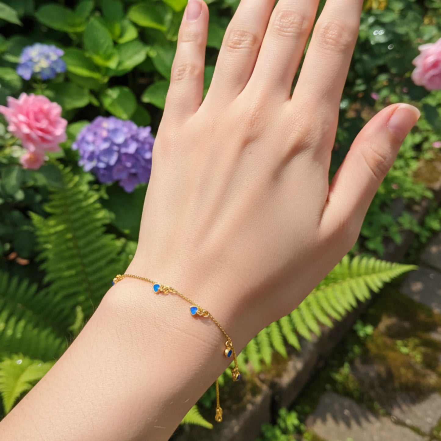 Hand wearing a gold plated bracelet with ocean blue heart charms with flowers in the background