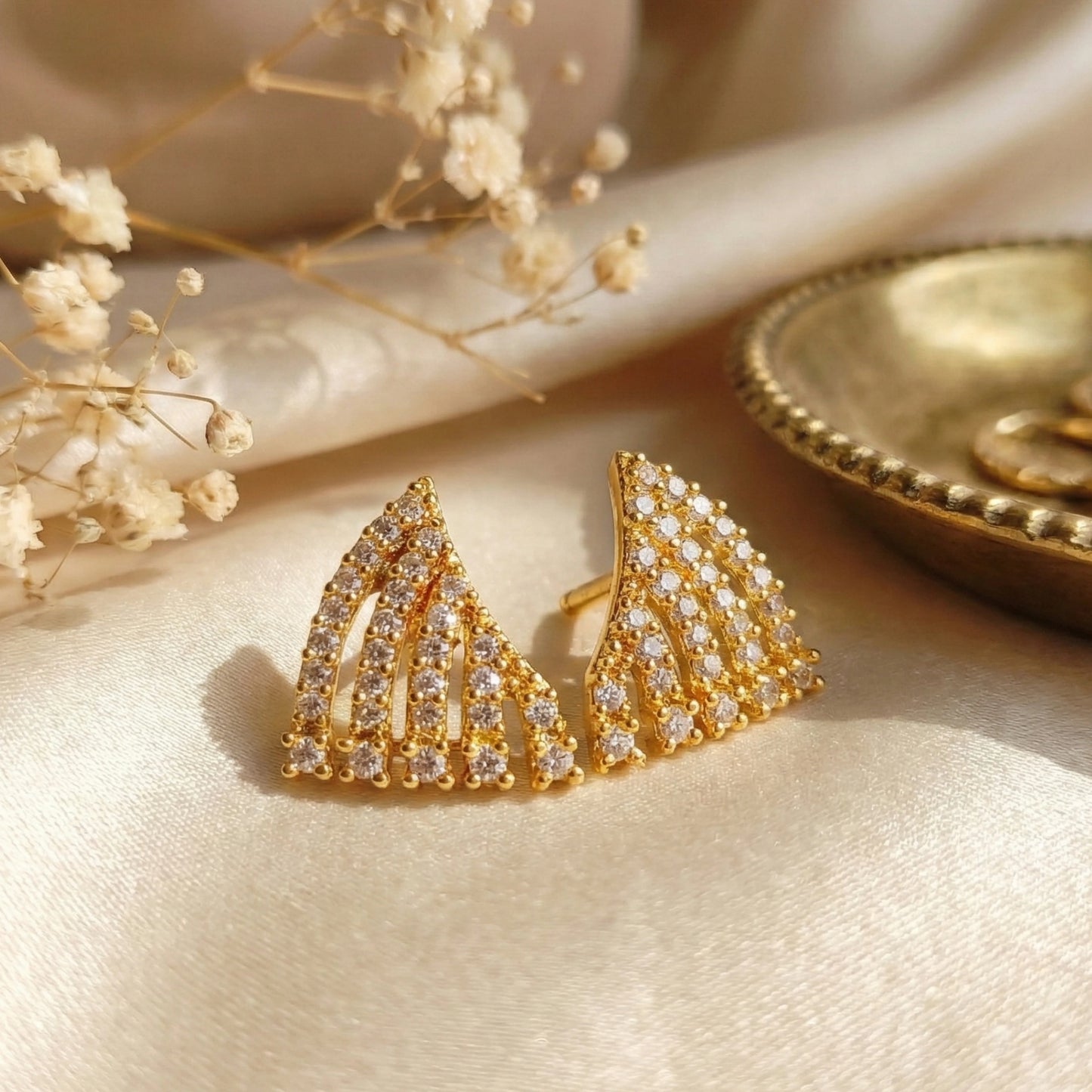 Gold plated earrings with AD accents on a beige surface with dried flowers.