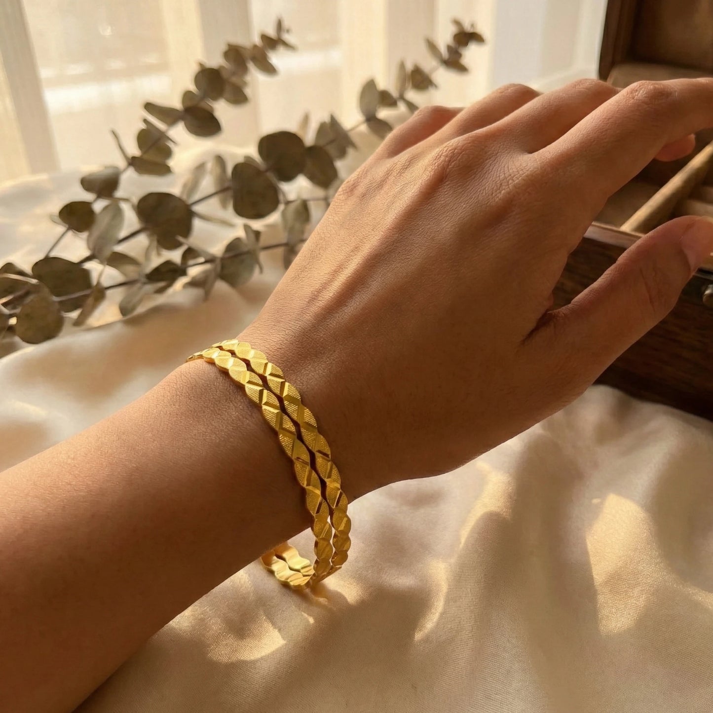 Hand wearing a gold plated braided bangles on a light fabric background with green leaves.