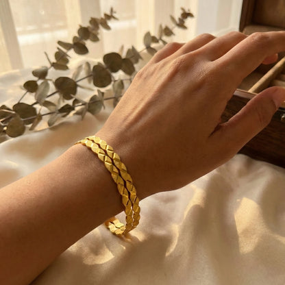 Hand wearing a gold plated braided bangles on a light fabric background with green leaves.