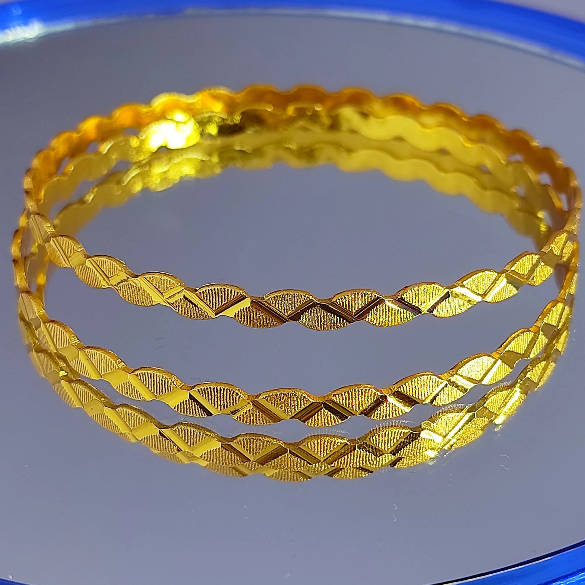 Two gold plated bangles with textured patterns on a reflective surface.