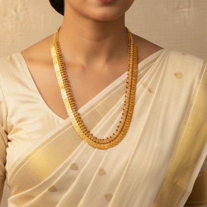 Woman wearing a white saree with gold patterns and a matching necklace.