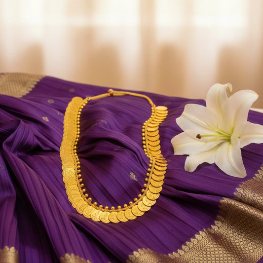 Gold plated kasu/coin haram necklace on a purple fabric with a white flower