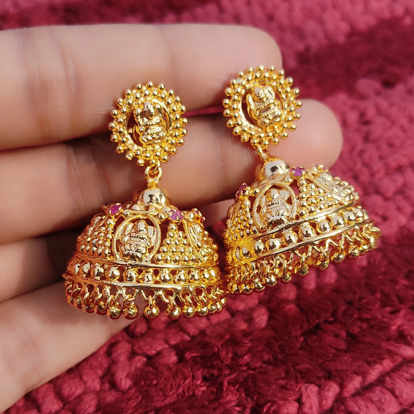 Gold plated Traditional Lakshmi Heritage Jimikki/Jhumka earrings held between fingers against a red textured background