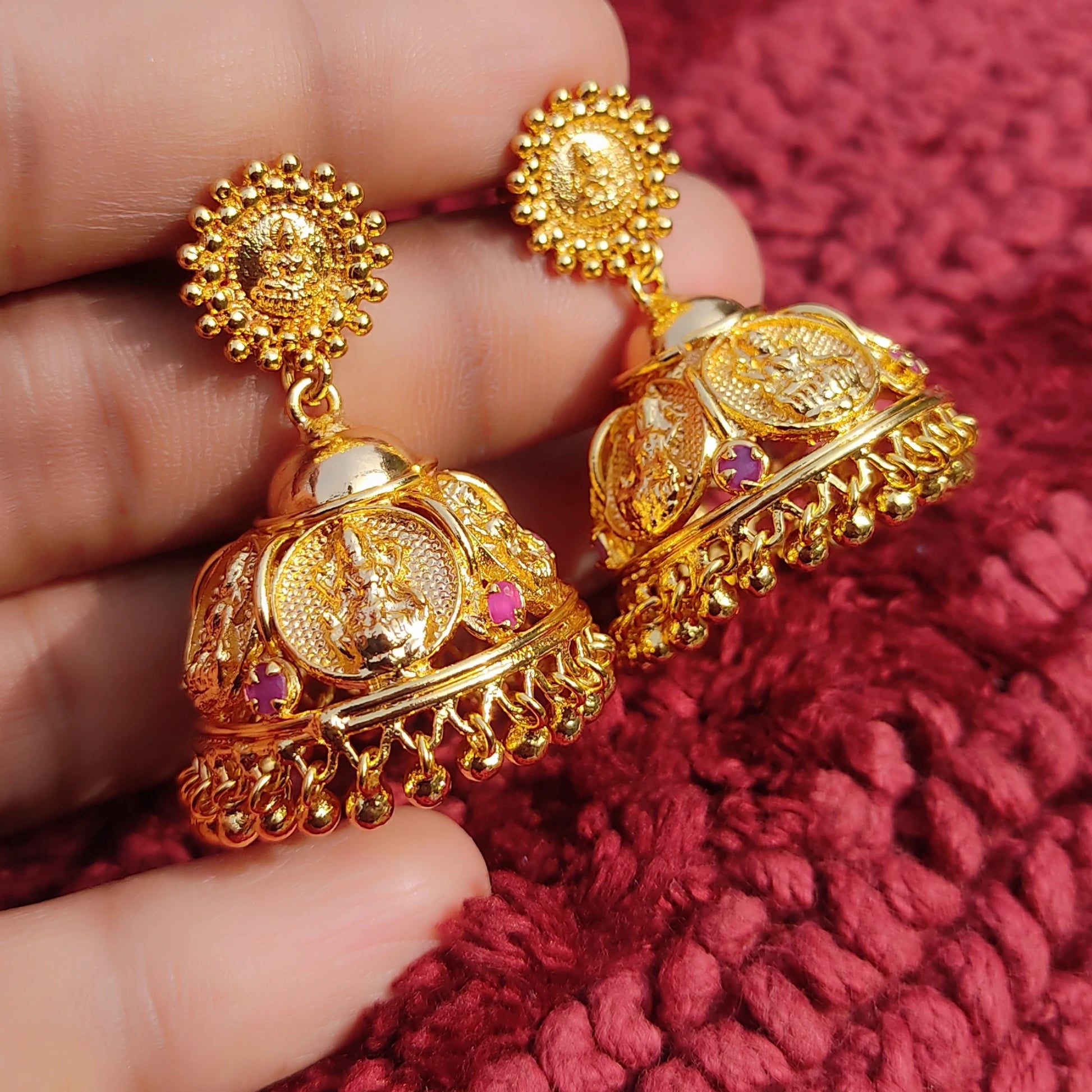 Gold plated traditional Lakshmi Temple Jimikki/Jhumkas earrings held between fingers against a red textured background