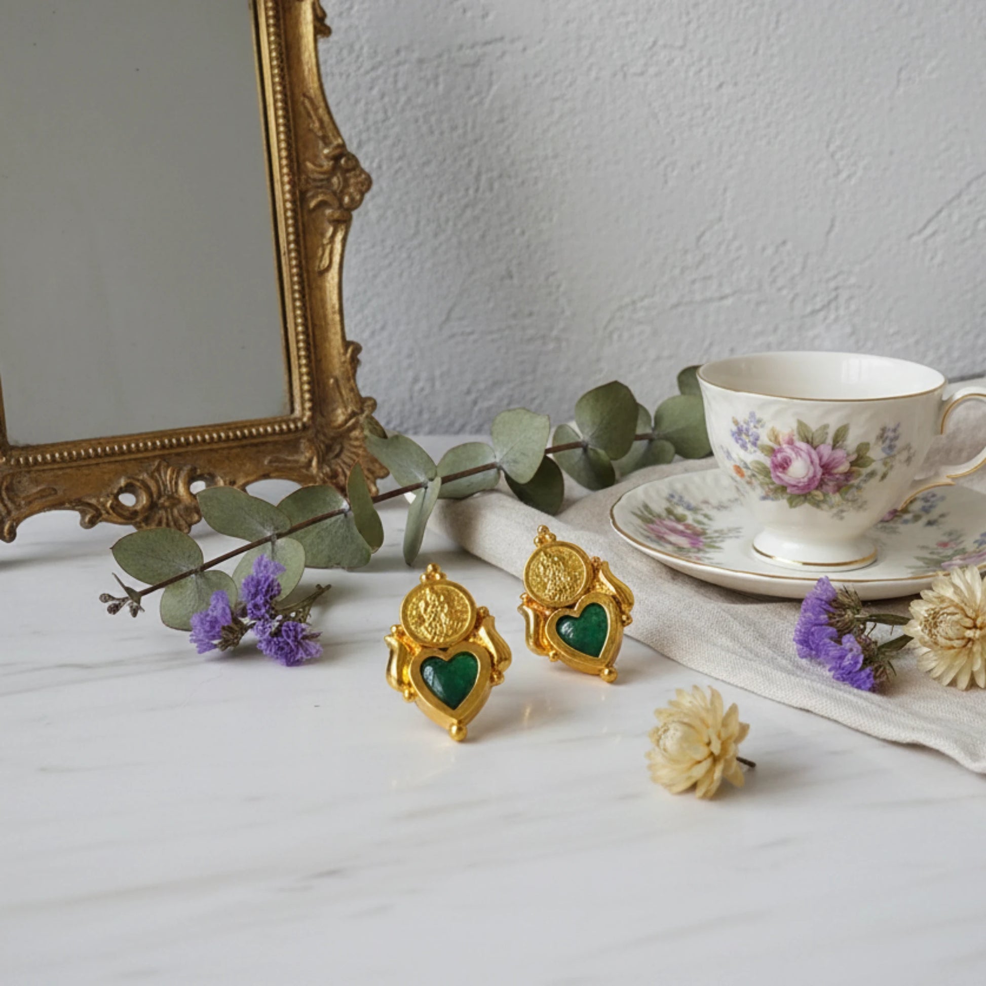 Gold plated palakka heart earrings with green inlay on a table with a teacup and decorative mirror.