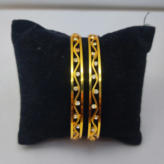 Two gold plated bangles with intricate designs on a black cushion.