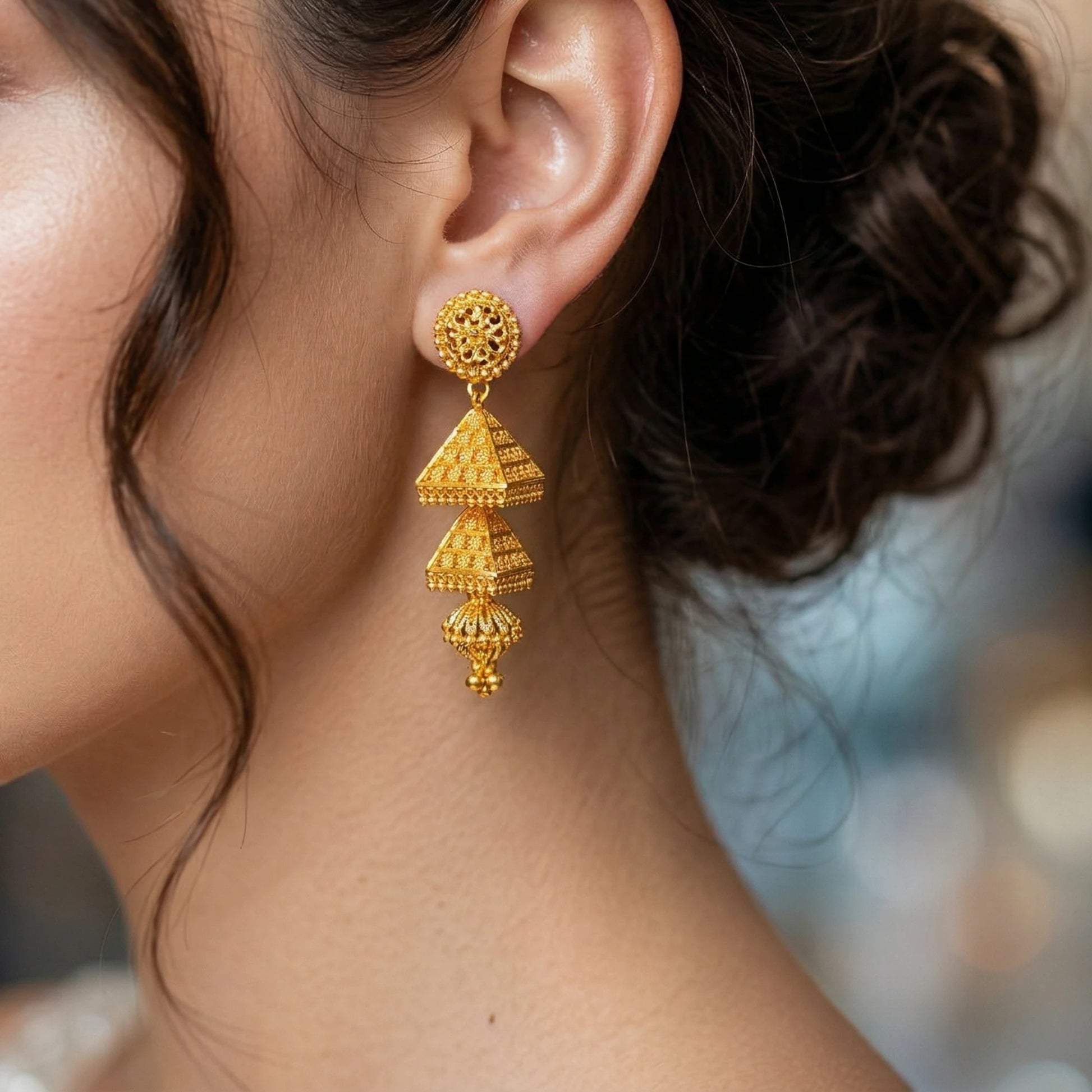 Close-up of a person wearing gold plated 3 tiered Jhumka/Jimikki earrings with a blurred background