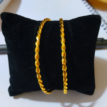 Two gold-plated twisted bangles displayed on a black velvet jewelry cushion against a light background.