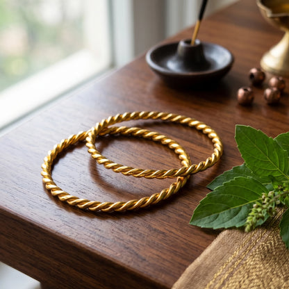 Two gold-plated twisted rope bangles displayed on a dark wooden table, with a small black dish, brass bells, green leaves, and a woven cloth nearby.