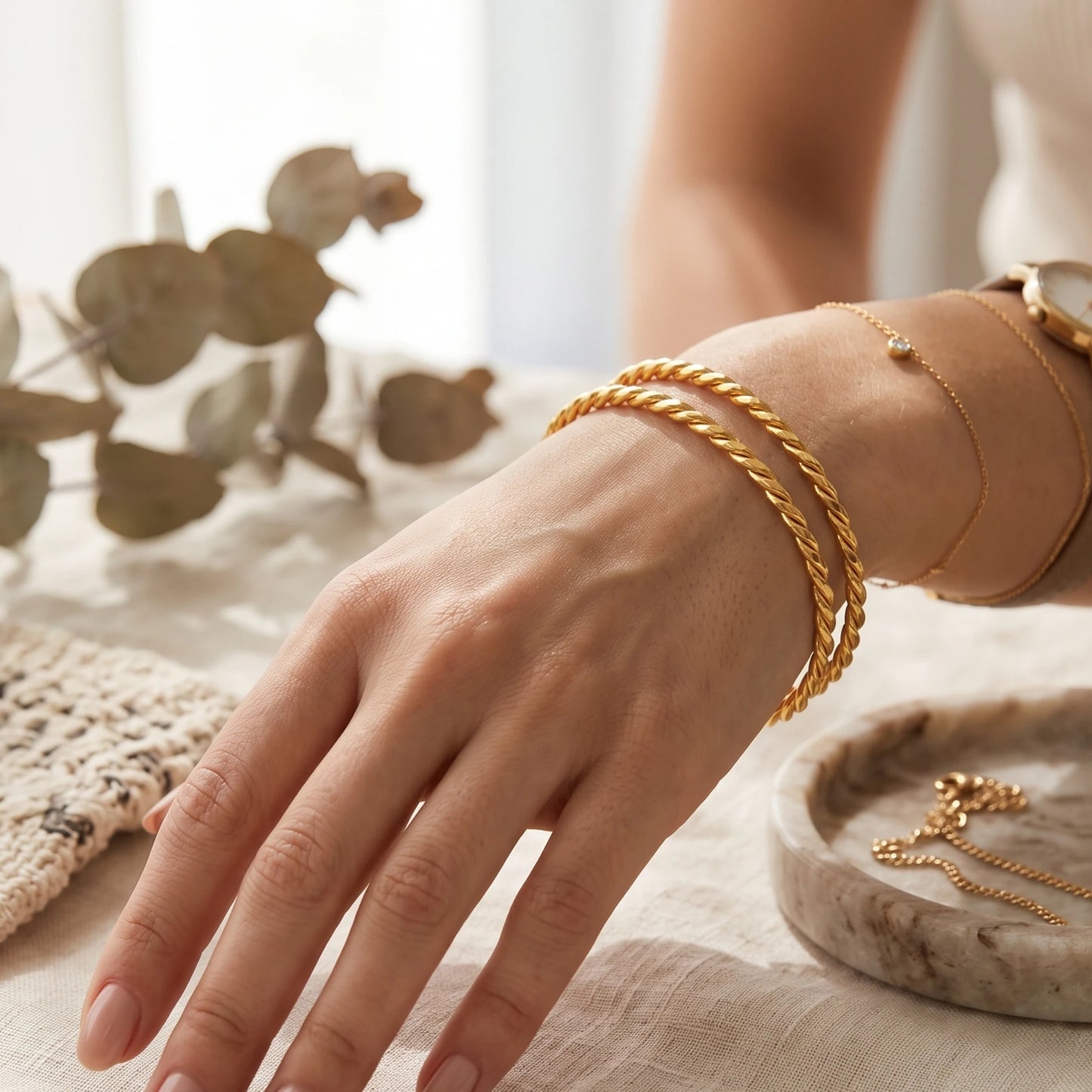 A close-up of a person's hand and wrist wearing multiple delicate gold plated bangles featuring a thick twisted rope-style, styled on a light fabric surface with eucalyptus leaves and a marble tray.