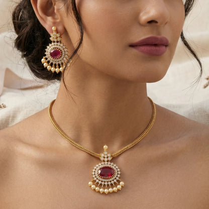 Close-up of a woman wearing a gold toned necklace with a red gemstone and matching earrings.