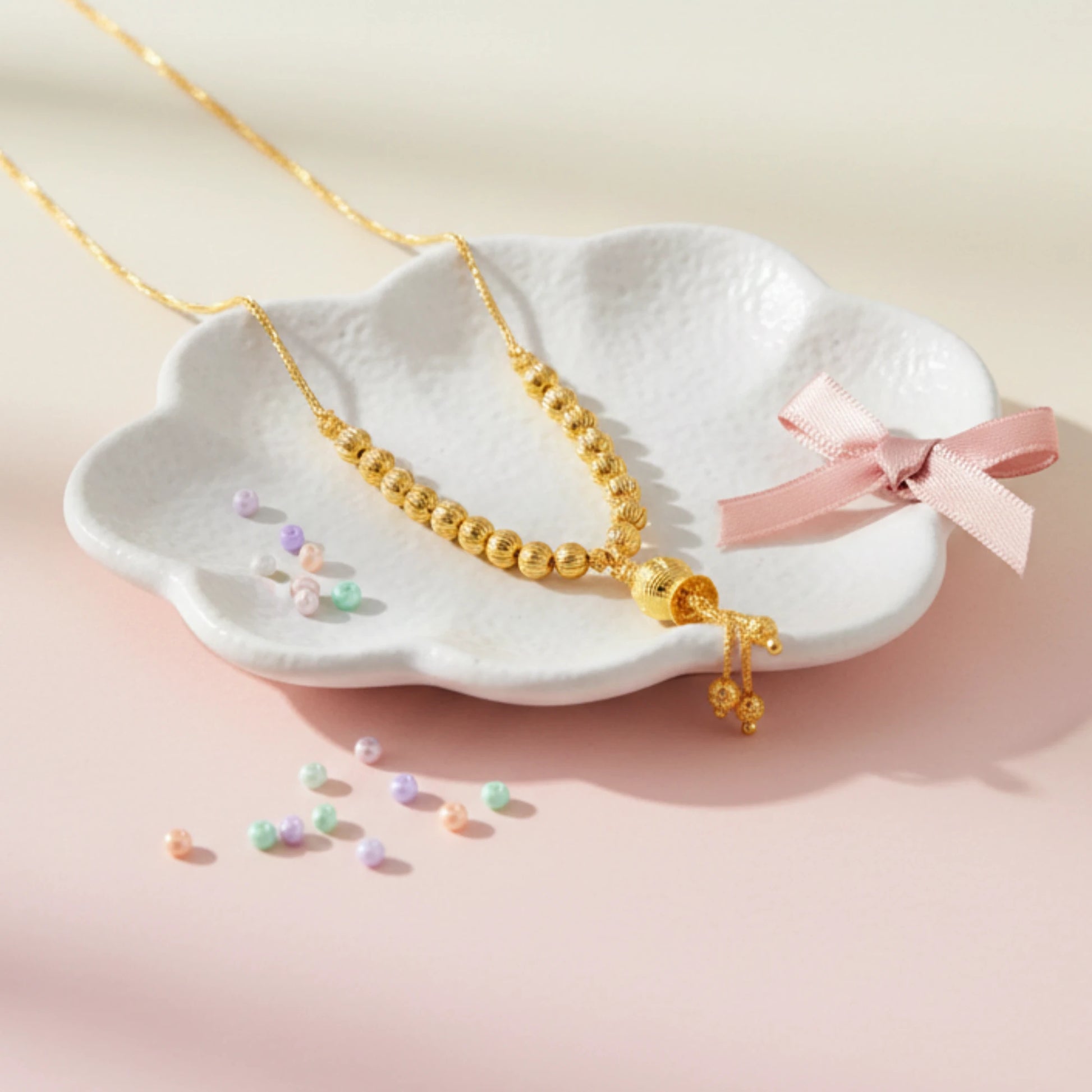Gold plated beaded necklace on a white ceramic dish with a pink ribbon against a light pink background
