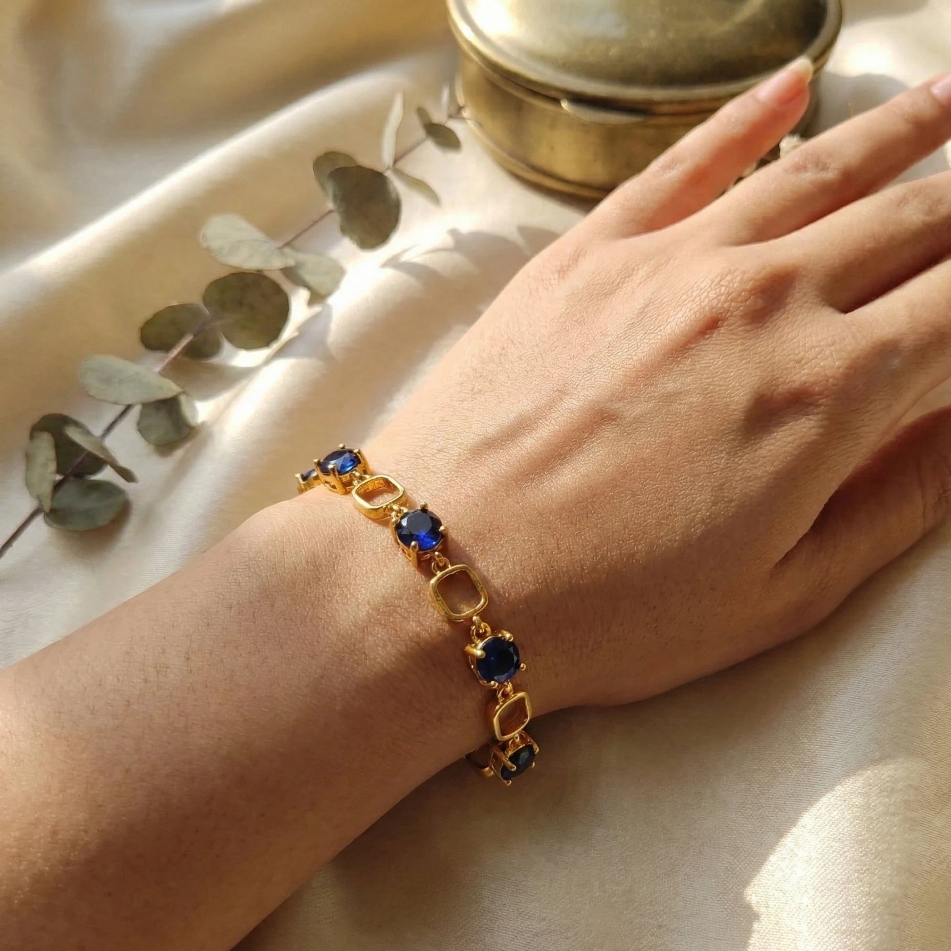 Gold plated bracelet with blue stones on a hand, placed on a light fabric background with eucalyptus leaves.