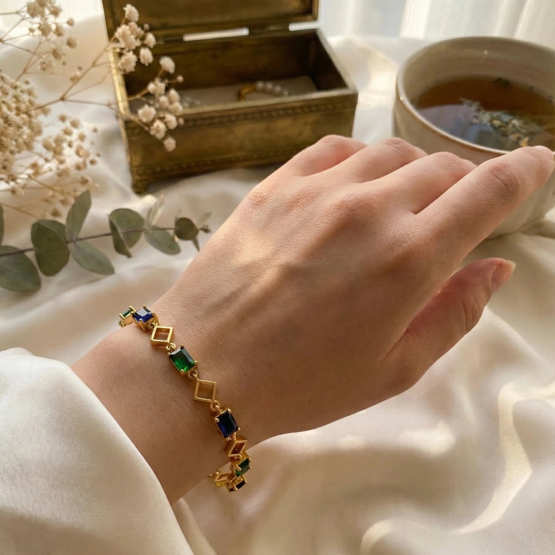 Hand wearing a gold plated bracelet with colored stones, next to a teacup and jewelry box on a soft surface.