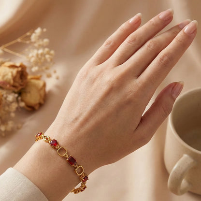 Hand wearing a gold plated bracelet with red stones, set against a soft background with a cup and flowers.