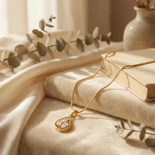Gold plated neckchain with a teardrop pendant on a soft surface with a book and leaves in the background