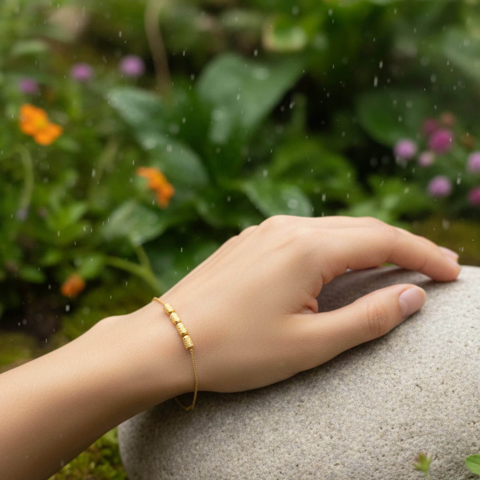 Hand wearing a gold plated bracelet on a stone with a natural background of greenery and flowers.