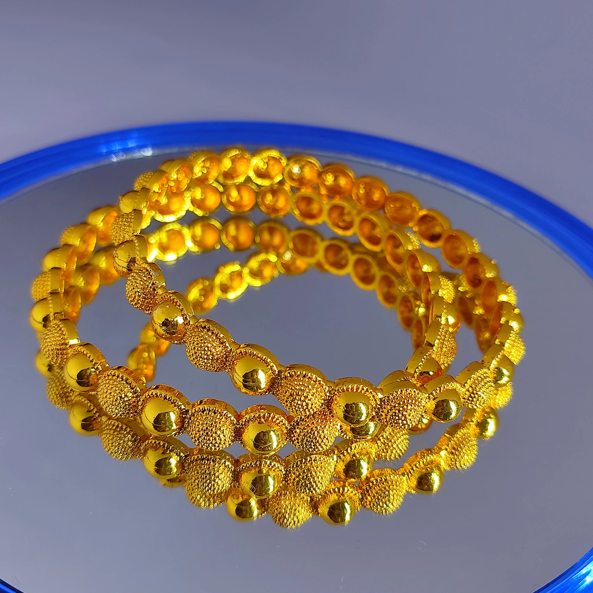 Two gold plated beaded bangles on a reflective surface with a blue rim.