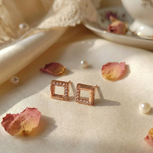 Rose gold square earrings on a textured surface with dried petals and pearls.