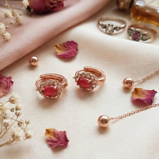 Rose gold jewelry with red gemstones on a light fabric background with dried flowers.