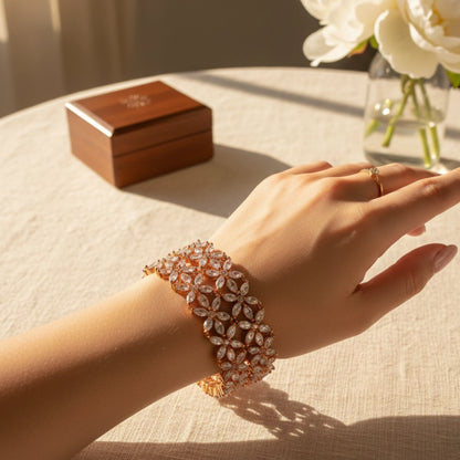 Hand wearing a sparkling Rose Gold Pair of bangles in floral design on a table with a wooden box and flowers in the background