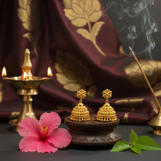 Gold plated earrings on a wooden stand with a pink flower and incense sticks in front of a dark fabric background.