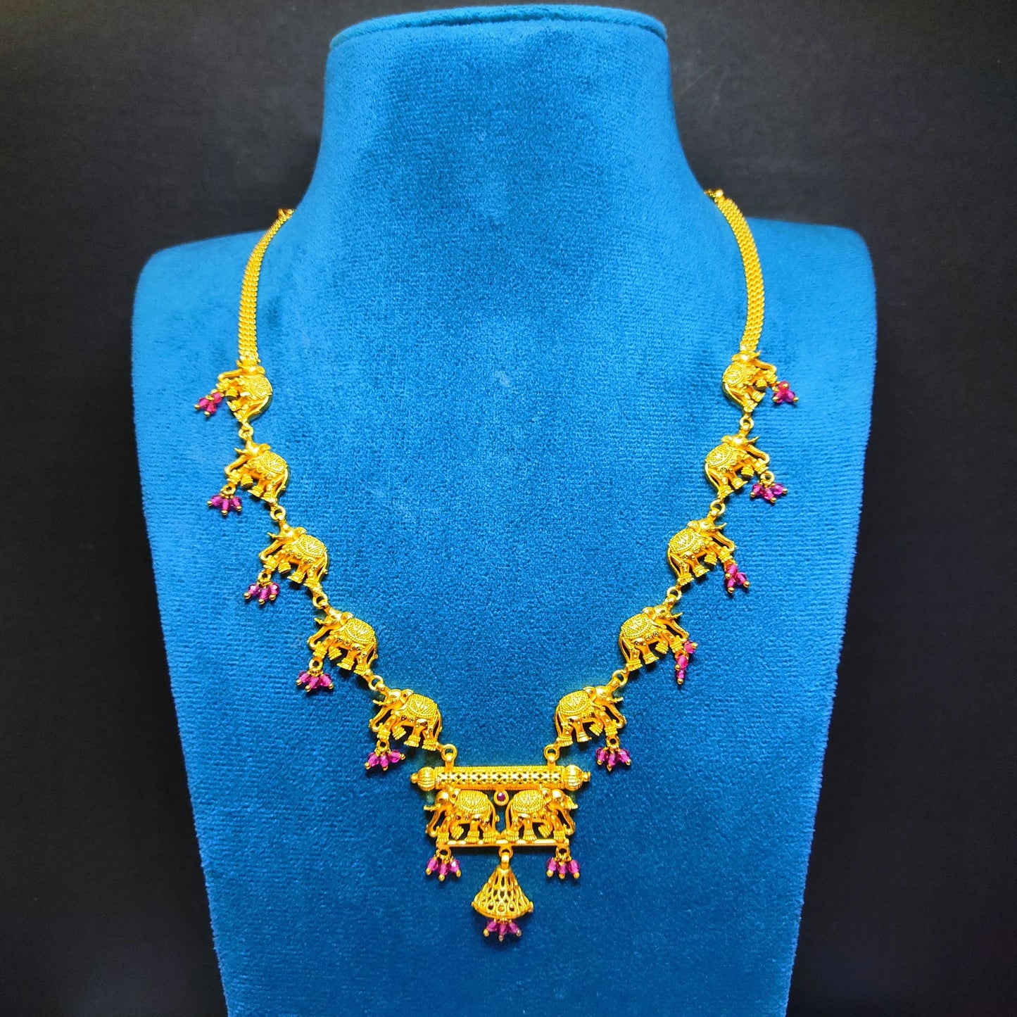 Gold plated necklace with elephant designs on a blue velvet stand against a black background