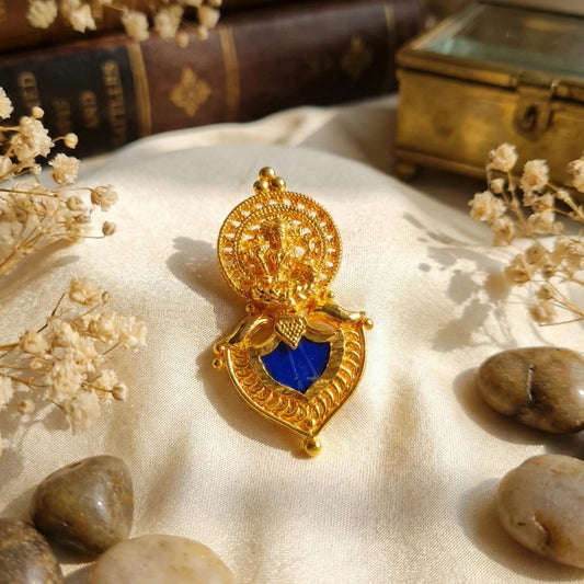 Gold plated heart-shaped pendant with blue palakka on a textured surface with dried flowers and stones.