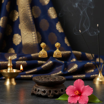 Gold plated earrings on a dark surface with a pink flower, incense sticks, and a blue fabric background.