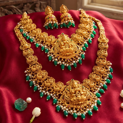 Table highlighting 1 long haram, 1 short haram with matching earrings, the design of the harams has goddess lakshmi as the pendant in the center and peacock design on both the sides of the necklace. the embellishments includes different kinds of stones, pearls and green embellishments and are targeted at occasion like weddings, grahapraveshams and festivities