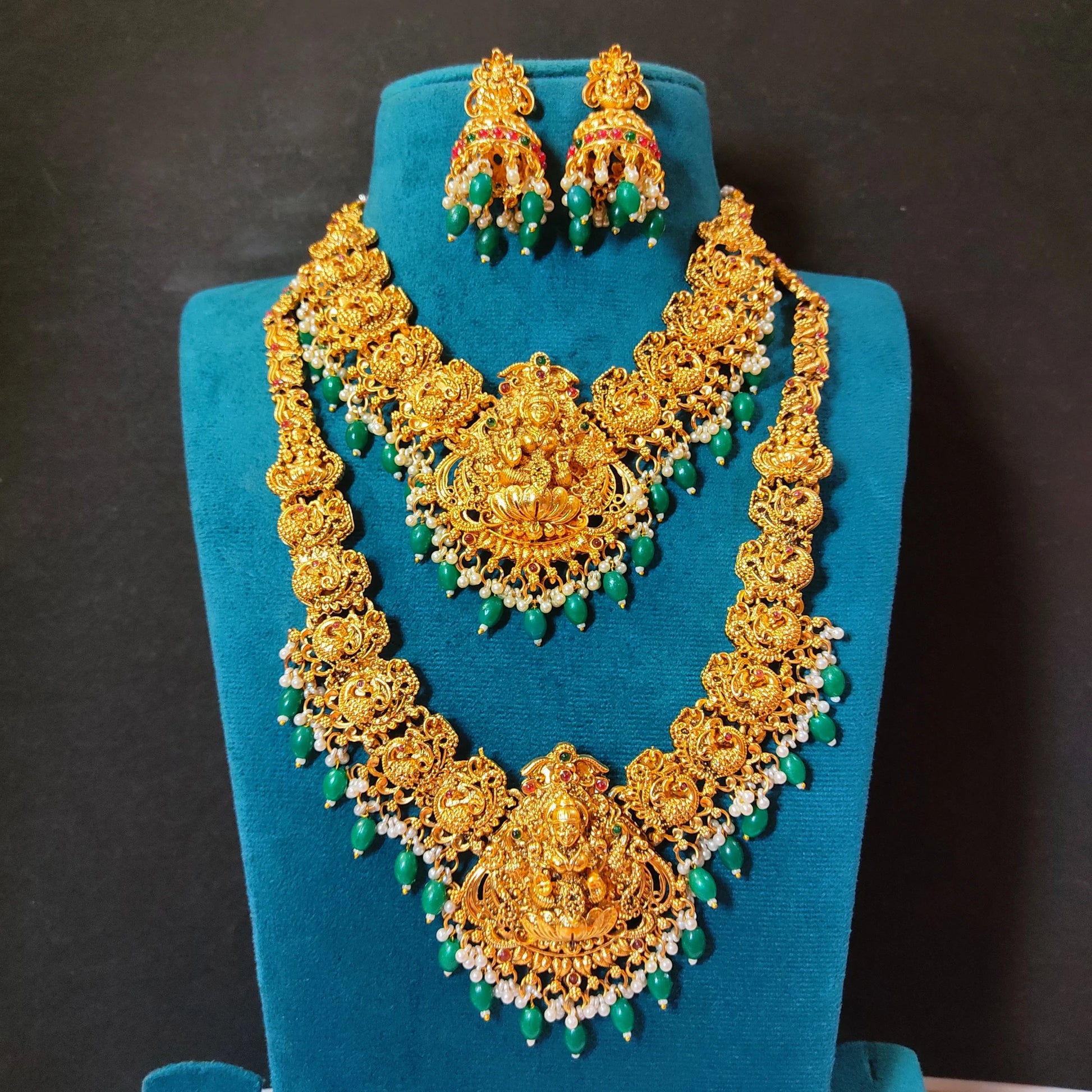 Mannequin highlighting 1 long haram, 1 short haram with matching earrings, the design of the harams has goddess lakshmi as the pendant in the center and peacock design on both the sides of the necklace. the embellishments includes different kinds of stones, pearls and green embellishments and are targeted at occasion like weddings, grahapraveshams and festivities