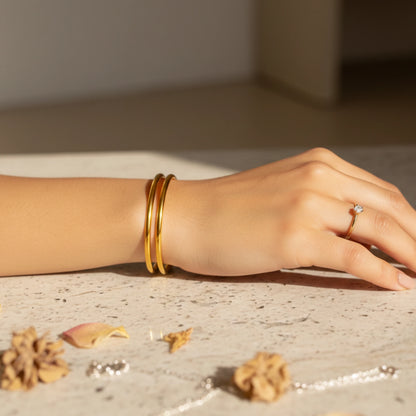 Hand wearing gold plated minimalist stackable bangles with a ring, on a textured surface with dried flowers.
