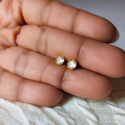 Gold plated second stud earrings with clear stones held between fingers on a white fabric background