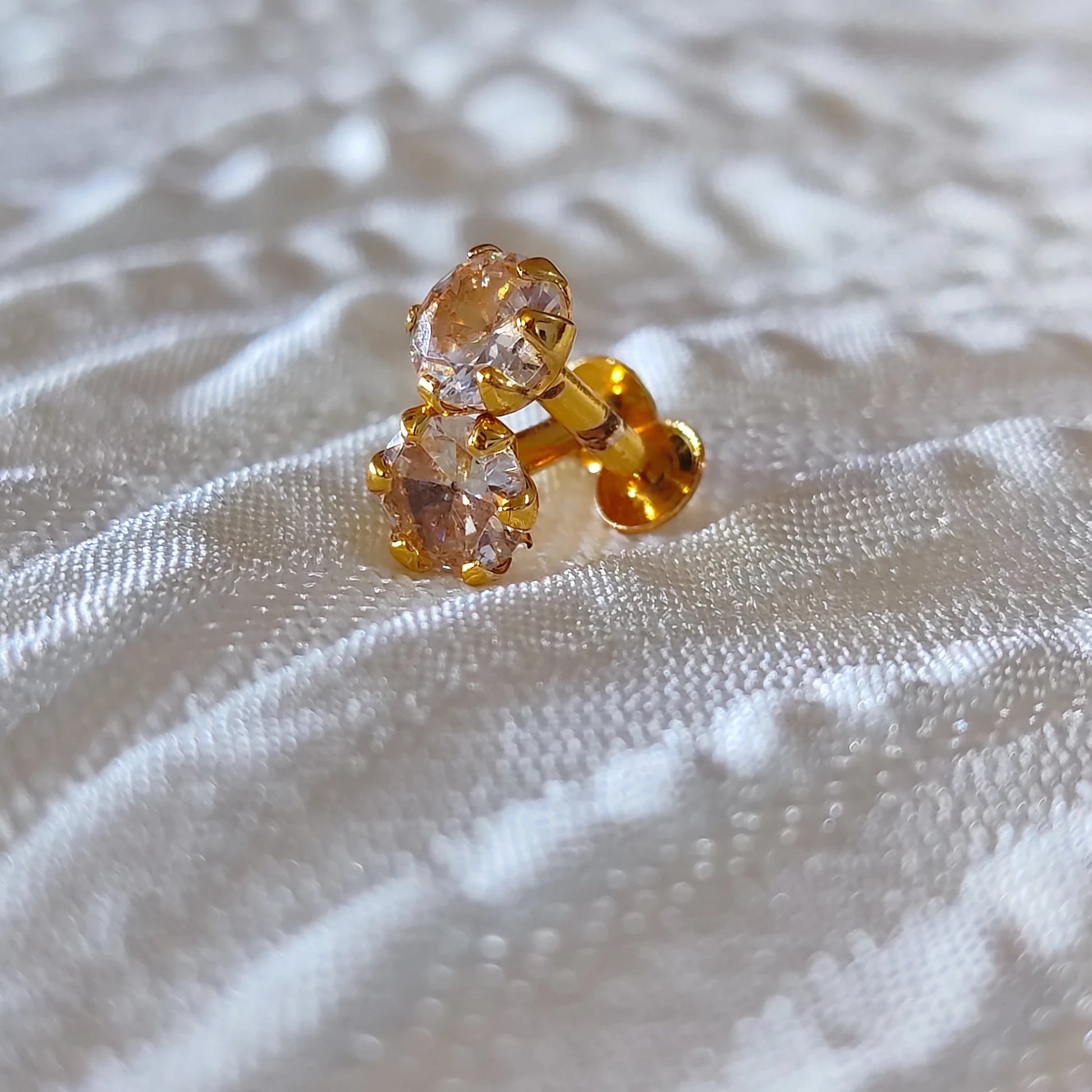 Gold plated second stud earrings with clear gemstones on a textured white fabric background