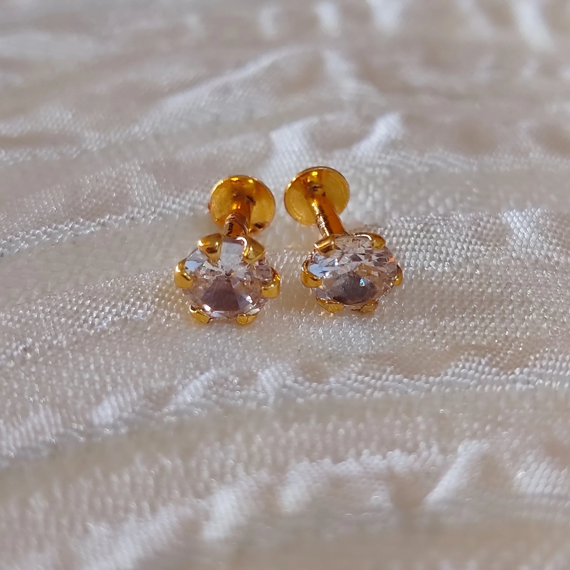 Gold plated second stud earrings with clear stones on a textured beige fabric background