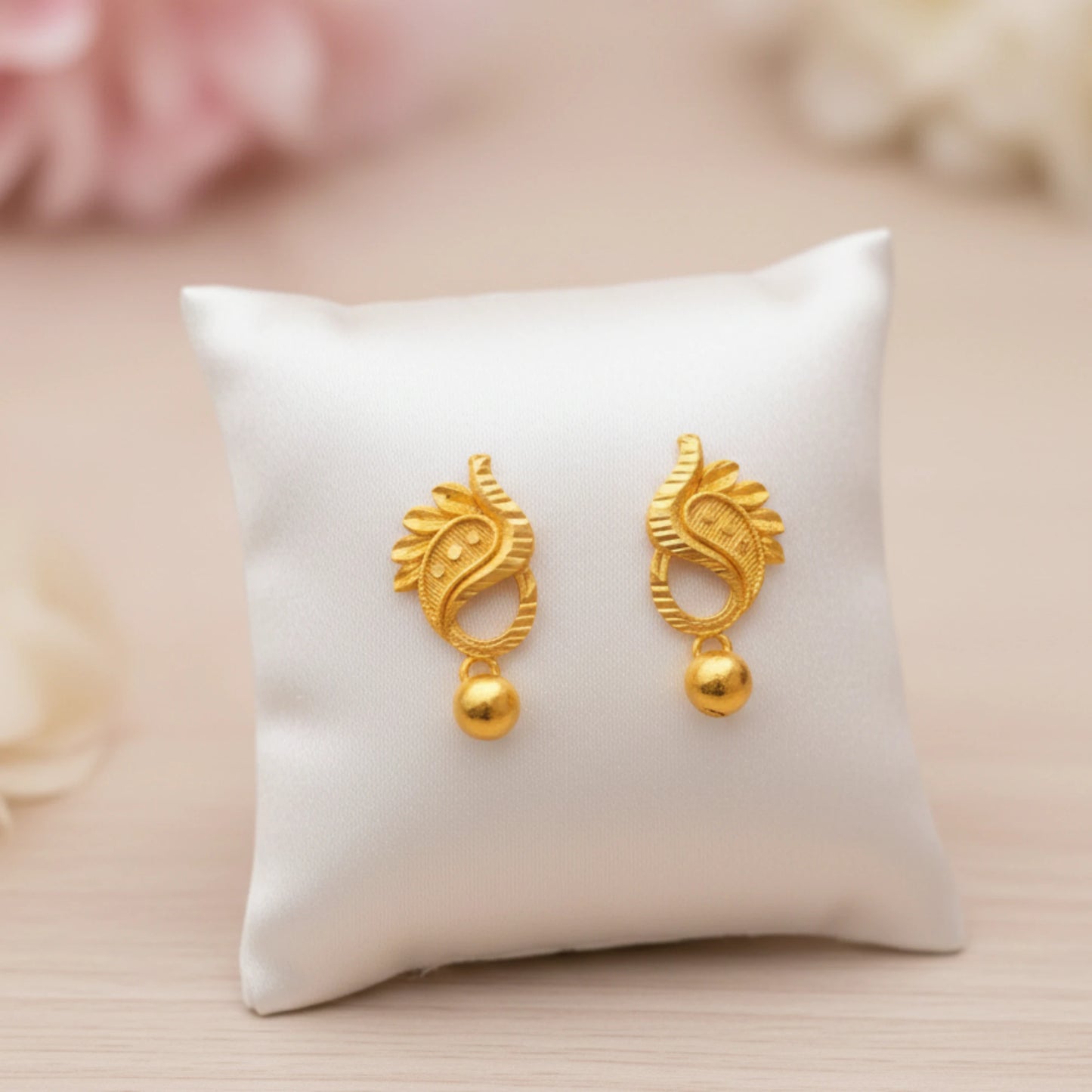 Gold plated manga mani earrings on a white cushion with a soft pink background