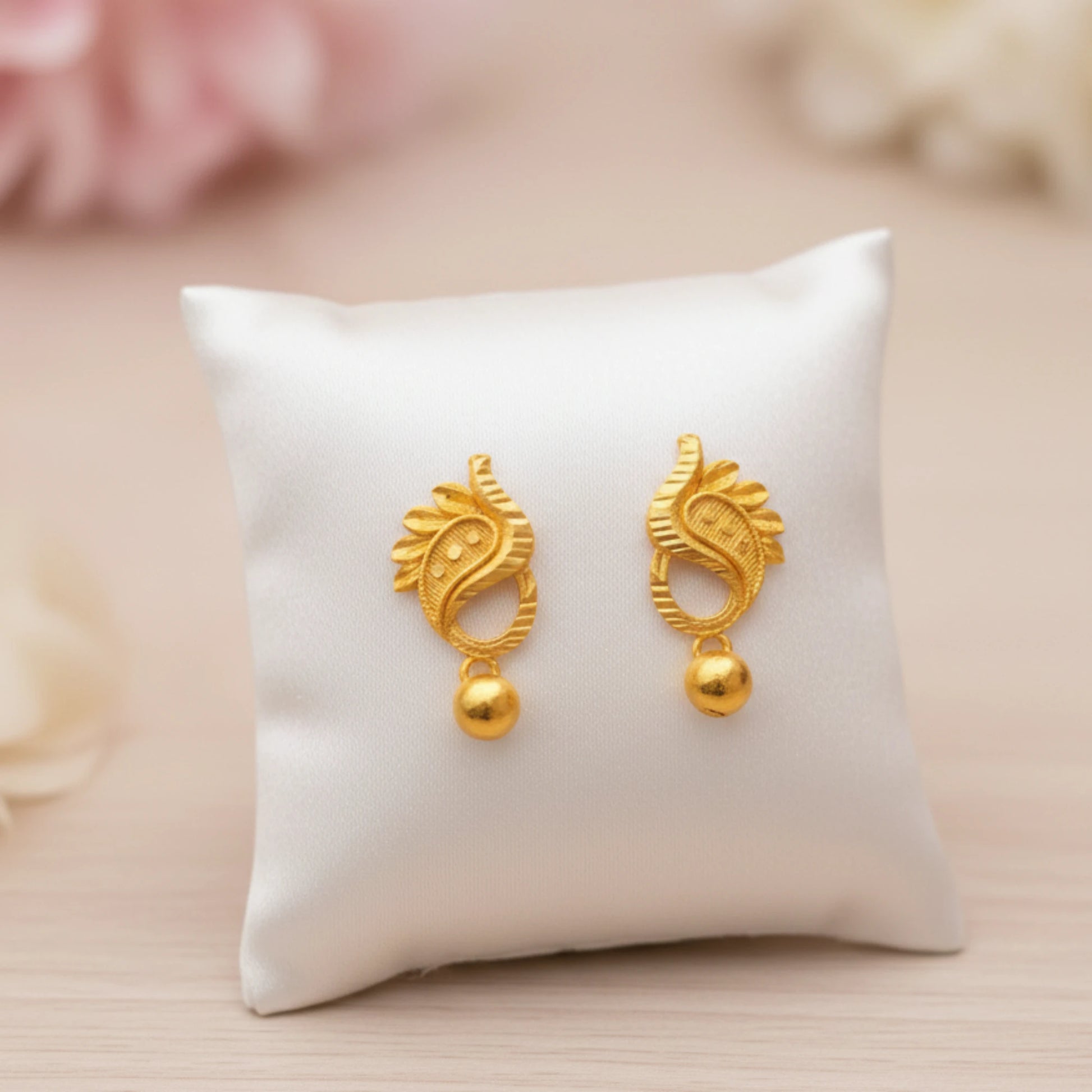 Gold plated manga mani earrings on a white cushion with a soft pink background