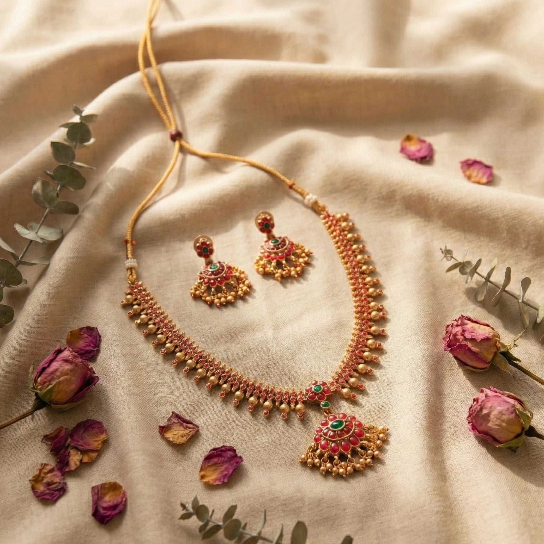 Ornate necklace and earrings on a beige fabric background with dried flowers and leaves.