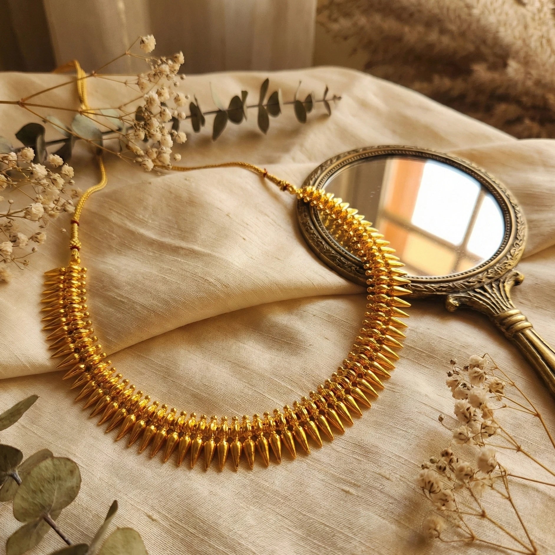 Gold plated Mulla Mottu necklace on a textured surface with dried flowers and a small mirror.